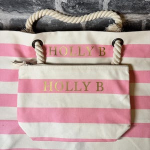 May include: A pink and white striped tote bag with a rope handle and a smaller zippered pouch. The bag has the name "HOLLY B" printed in gold on the front.