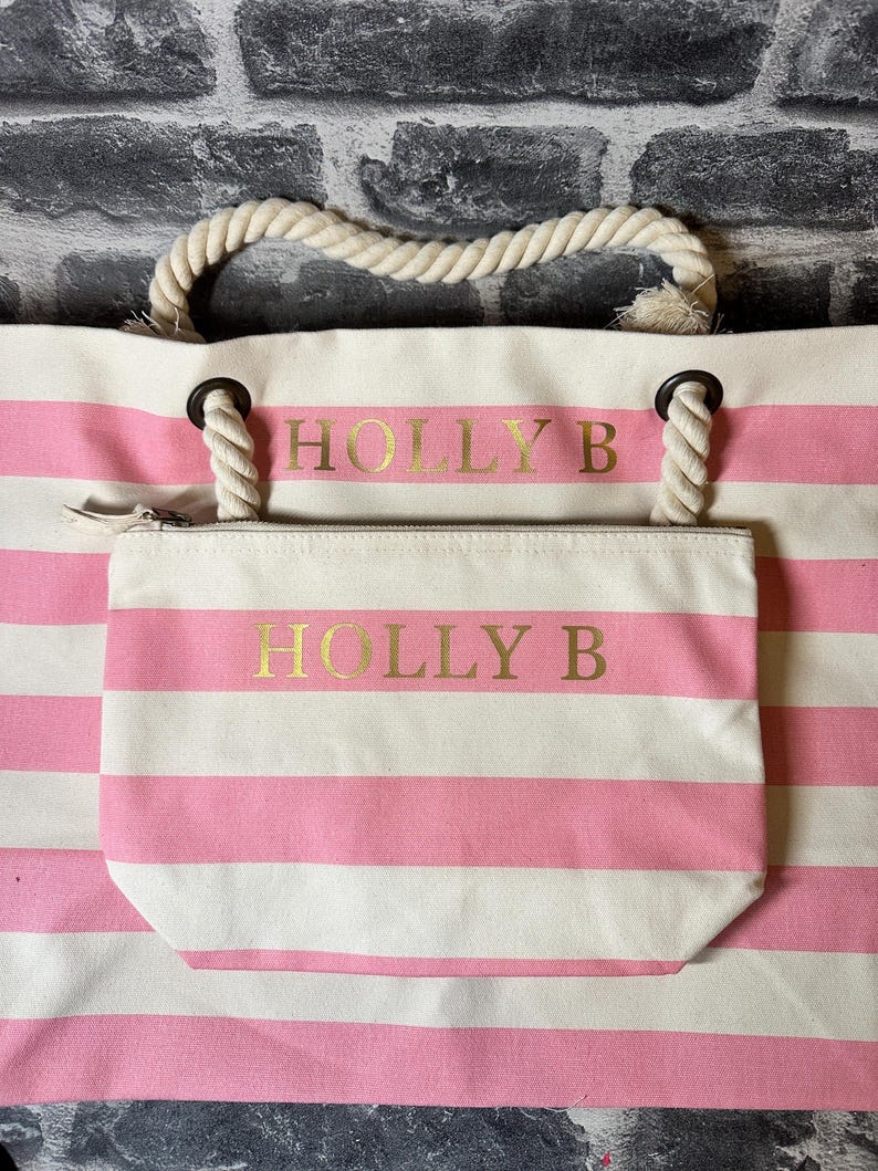 May include: A pink and white striped tote bag with a rope handle and a smaller zippered pouch. The bag has the name "HOLLY B" printed in gold on the front.