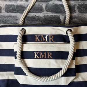 May include: A white and navy blue striped tote bag with rope handles. The bag has a zippered pocket on the front with the initials "KMR" embroidered in gold on both the bag and the pocket.