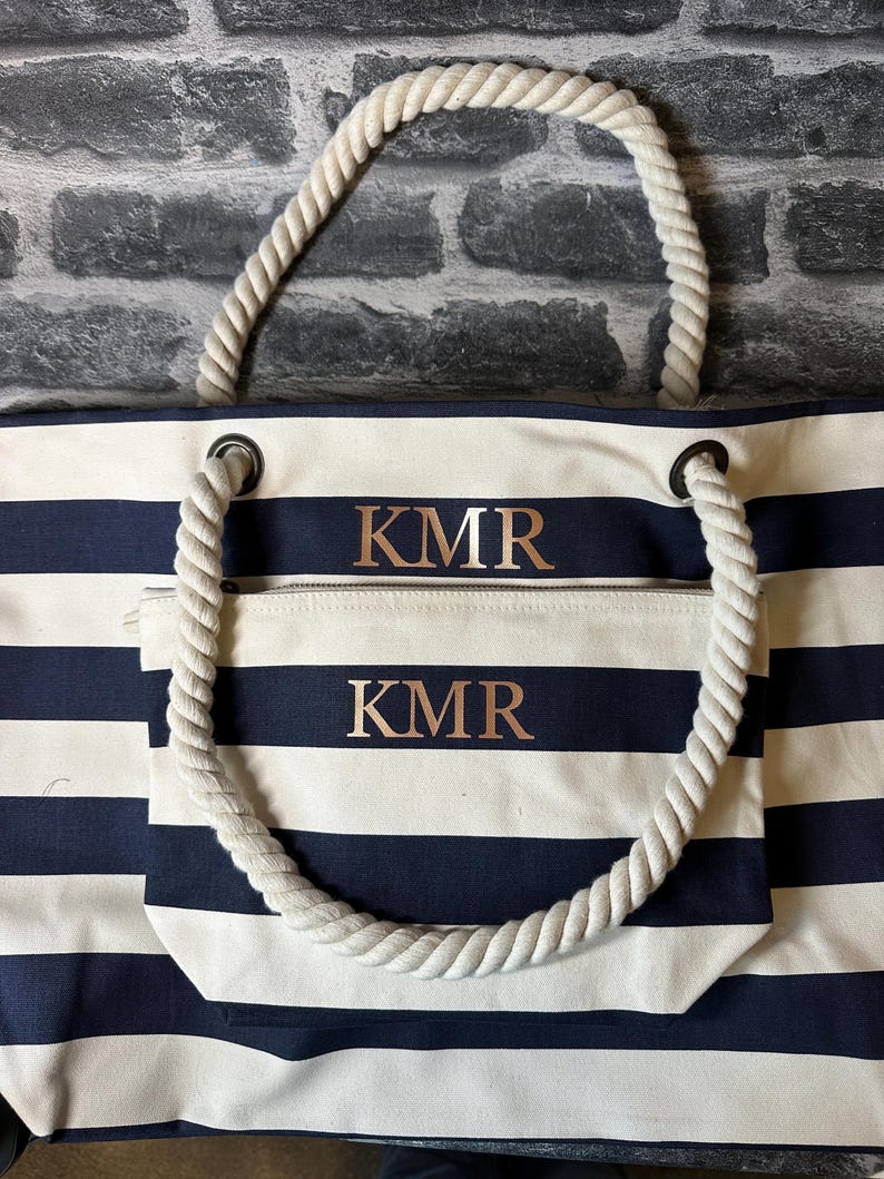 May include: A white and navy blue striped tote bag with rope handles. The bag has a zippered pocket on the front with the initials "KMR" embroidered in gold on both the bag and the pocket.