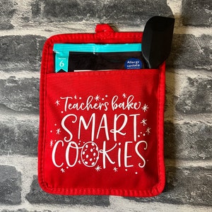 May include: Red oven mitt with white text that says "Teachers bake SMART COOKIES". The oven mitt has a pocket with a blue packet of cookie mix and a black spatula.