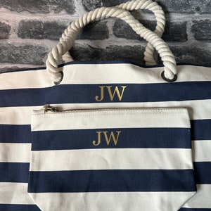 A white and navy blue striped tote bag with rope handles. The bag has a zippered pocket on the front with the initials 'JW' embroidered in gold on both the bag and the pocket.