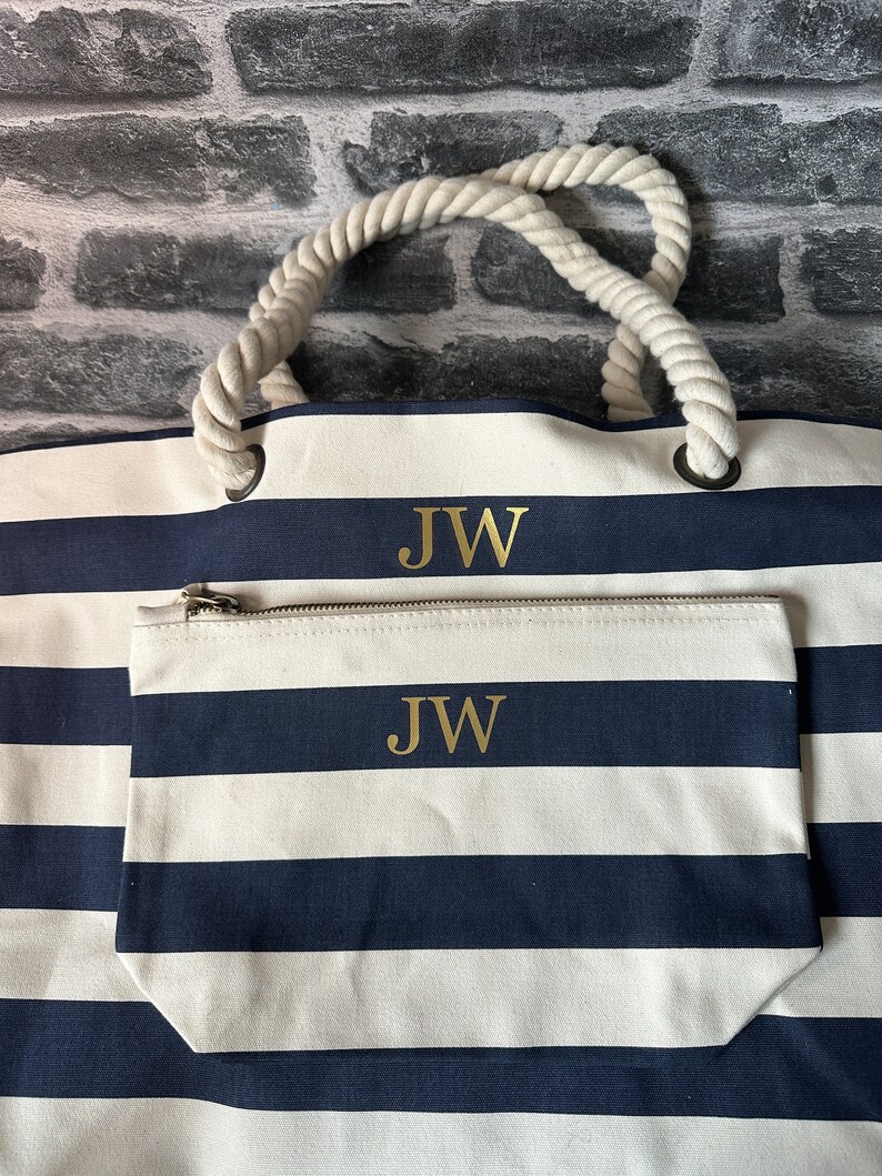 A white and navy blue striped tote bag with rope handles. The bag has a zippered pocket on the front with the initials 'JW' embroidered in gold on both the bag and the pocket.