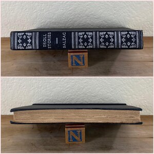 May include: A hardcover book titled "Droll Stories" by Balzac, with a dark blue cover and decorative white and silver patterns. The book is propped up by a wooden block with the letter "N" on it.