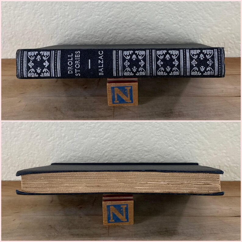 May include: A hardcover book titled "Droll Stories" by Balzac, with a dark blue cover and decorative white and silver patterns. The book is propped up by a wooden block with the letter "N" on it.