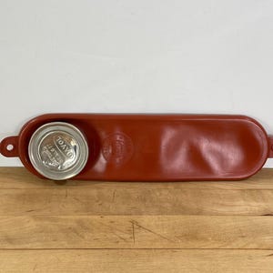 May include: A rust-colored, oval-shaped tray with a circular indentation holding a silver can. The tray has a raised edge and two small holes on either end. The can has a silver lid with white text.