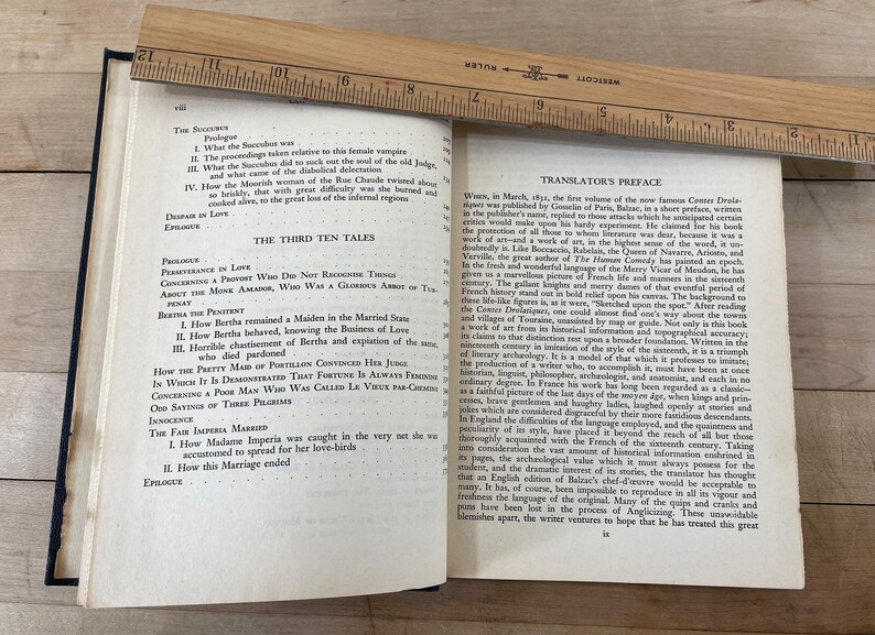 May include: An open book with text, a wooden ruler, and a dark blue cover. The ruler, marked in inches, rests across the pages. The text includes titles like "The Third Ten Tales" and "Translator's Preface."