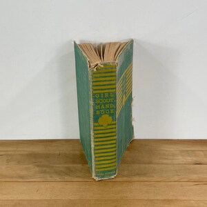 May include: Vintage Girl Scout Handbook with a teal cover and yellow stripes. The book's spine displays the title "GIRL SCOUT HAND BOOK" in yellow lettering. The Girl Scout trefoil emblem is also visible. The book is aged with visible wear.