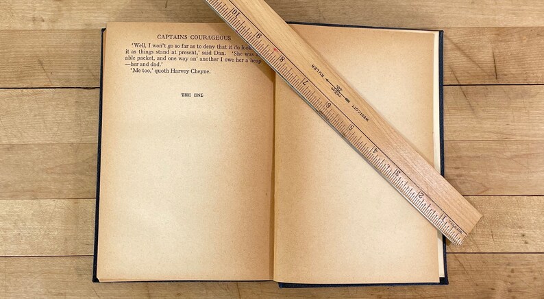 May include: An open book with aged, yellowed pages, titled "CAPTAINS COURAGEOUS." A wooden ruler, marked in inches, rests diagonally across the pages. The text "THE END" is visible.