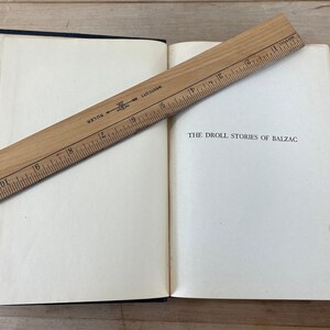 May include: An open book with the title "THE DROLL STORIES OF BALZAC" is shown with a wooden ruler across the pages. The book has a dark blue cover and off-white pages. The ruler is marked with inches.