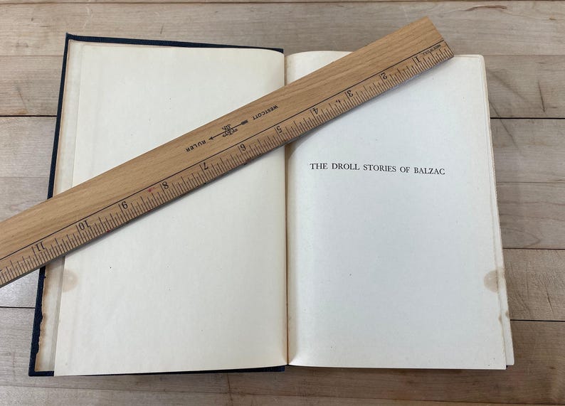 May include: An open book with the title "THE DROLL STORIES OF BALZAC" is shown with a wooden ruler across the pages. The book has a dark blue cover and off-white pages. The ruler is marked with inches.