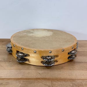 May include: A round tambourine with a light brown wooden frame and a natural skin drumhead. The frame has small metal jingles and is resting on a wooden surface. A musical instrument.