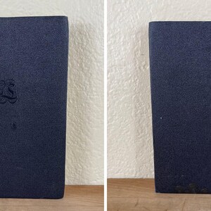 May include: Two images of a navy blue book with a textured cover. The front cover has an embossed design in the center. The book is standing upright on a wooden surface against a white wall.
