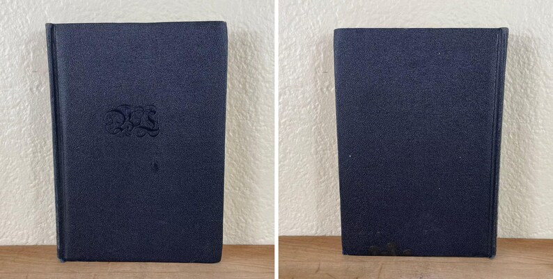 May include: Two images of a navy blue book with a textured cover. The front cover has an embossed design in the center. The book is standing upright on a wooden surface against a white wall.
