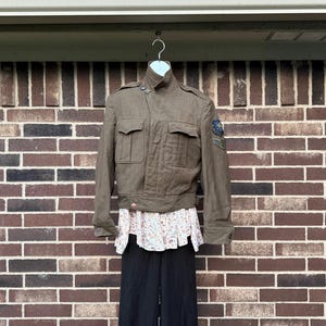 May include: A brown military-style jacket with two chest pockets and a high collar. The jacket has patches on the sleeve and is displayed on a hanger. The jacket is paired with a floral top and black pants.