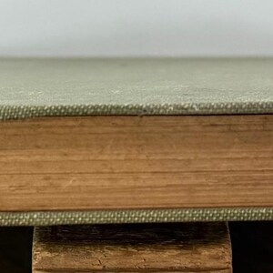 K&ouml;nnte beinhalten: Ein gebundenes Buch mit einem strukturierten, salbeigr&uuml;nen Stoffeinband und hellbraunen, gealterten Papierkanten. Das Buch liegt horizontal und zeigt den Buchr&uuml;cken sowie die Dicke der Seiten, die eine warme, hellbraune Vintage-F&auml;rbung aufweisen.