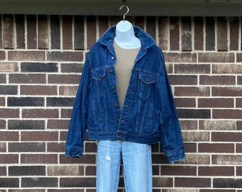 Puede incluir: Chaqueta vaquera azul oscuro con cuello y botones, combinada con un suéter beige y vaqueros rotos azul claro. El conjunto se exhibe en un maniquí blanco sobre un fondo de pared de ladrillo.