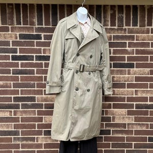 May include: A beige double-breasted trench coat with a matching waist belt, wide lapels, and shoulder epaulets. The coat is worn over a light-colored patterned shirt and black trousers, featuring dark buttons and adjustable sleeve straps.