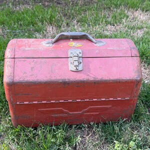 May include: A rectangular red metal toolbox with a weathered finish, featuring a central silver metal latch, a top handle, and a molded wing-like emblem on the front. The box has a hinged lid and shows signs of wear and surface oxidation throughout.