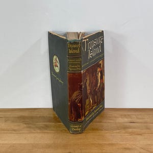 Könnte beinhalten: Ein Vintage-Exemplar von "Die Schatzinsel" von Robert Louis Stevenson. Das Buch hat einen dunkelgrünen Einband mit goldener Schrift und Illustrationen. Der Buchrücken zeigt den Titel und den Namen des Autors. Herausgegeben von Grosset & Dunlop.