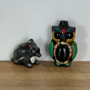 May include: Two ceramic figurines: a black squirrel with white accents and a colorful owl. The owl has green wings, yellow and black eyes, and red and white details. Both are on a wooden surface.