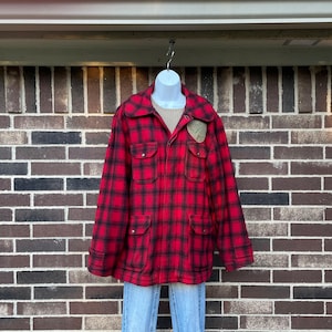 May include: A red and black plaid jacket featuring a large collar, four front pockets with button closures, and a tan patch on the upper left chest. The jacket is worn over a beige ribbed shirt and light blue denim jeans on a mannequin.
