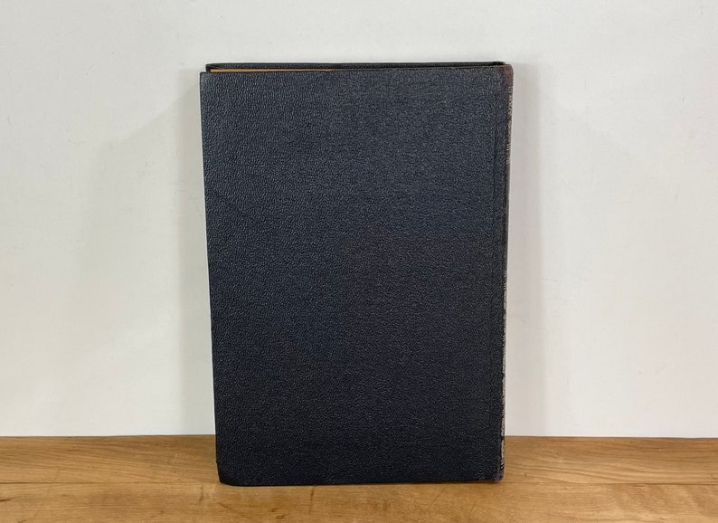 May include: A vintage book with a textured, dark blue cover. The spine shows signs of wear, with a slightly faded appearance. The book is standing upright on a wooden surface, suggesting it's part of a collection or display.
