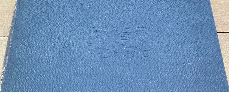 May include: A close-up of a blue leather-bound book. The cover has a textured surface and an embossed design in the center. The book's edges are visible, showing the pages and binding.