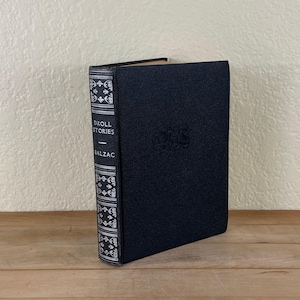 May include: A hardcover book titled "Droll Stories" by Balzac. The book has a dark blue cover with embossed lettering and a decorative spine with white and silver accents. The book is standing upright on a wooden surface.