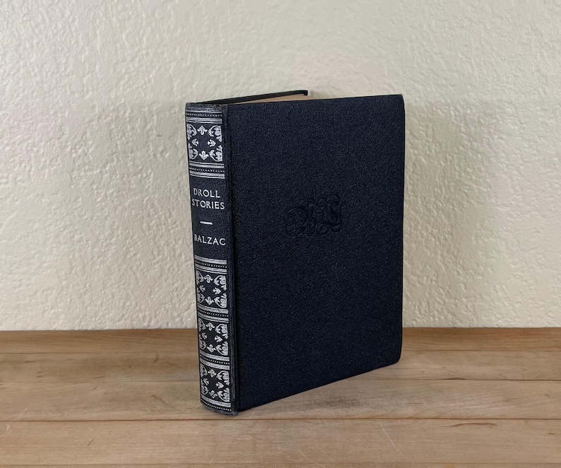 May include: A hardcover book titled "Droll Stories" by Balzac. The book has a dark blue cover with embossed lettering and a decorative spine with white and silver accents. The book is standing upright on a wooden surface.