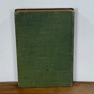 K&ouml;nnte beinhalten: Ein rechteckiges Hardcover-Buch mit einem strukturierten, olivgr&uuml;nen Stoffeinband. Das Buch ist vertikal aufgestellt und zeigt den Vorderdeckel mit sichtbaren Abnutzungserscheinungen an den Kanten und Ecken sowie einen dunklen Buchr&uuml;cken.