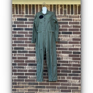 May include: An olive green flight suit featuring a front zipper closure, long sleeves, and multiple utility pockets on the legs and chest. A circular black patch is positioned on the right chest area, and the garment is displayed on a white mannequin.