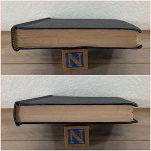 May include: A hardcover book with a dark blue cover and aged, tan-colored pages. The book is resting on a wooden block with the letter "N" in blue. The book's spine is visible, showing the thickness of the pages.