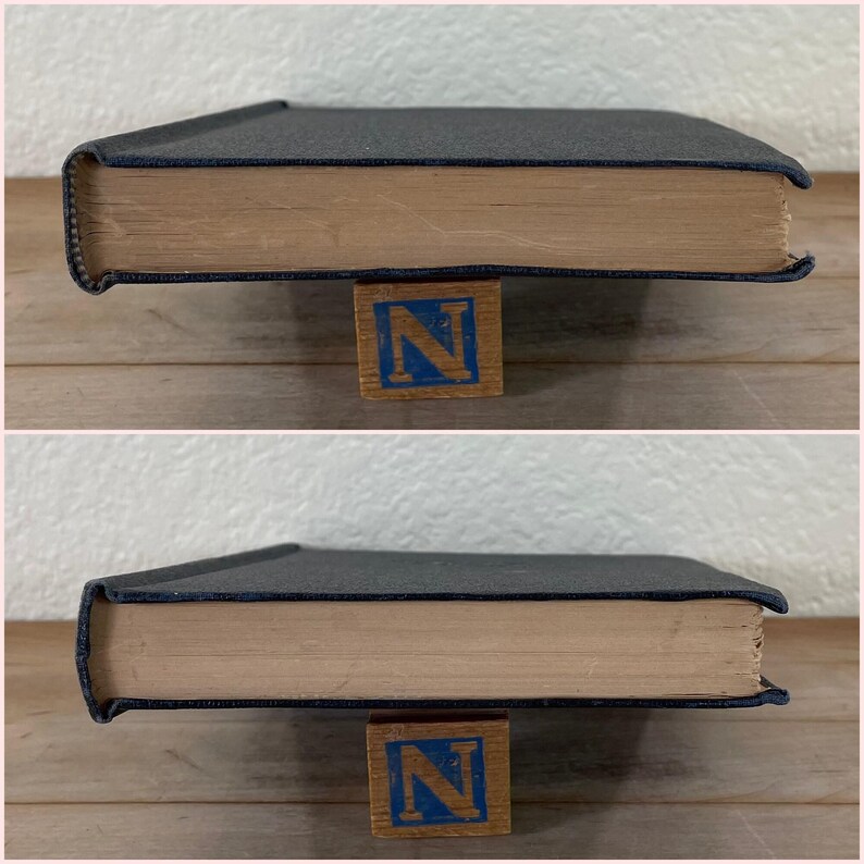 May include: A hardcover book with a dark blue cover and aged, tan-colored pages. The book is resting on a wooden block with the letter "N" in blue. The book's spine is visible, showing the thickness of the pages.