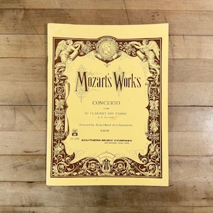 May include: A sheet music cover for Mozart's Works Concerto for B-flat Clarinet and Piano, K.V. No. 622, revised by Jean Albert de la Tournerie. The cover features a cream background with dark brown ornate borders, cherubs, and musical motifs.