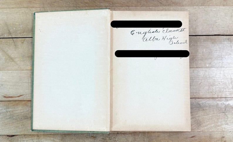 K&ouml;nnte beinhalten: Ein aufgeschlagenes Hardcover-Buch mit gr&uuml;nem Buchr&uuml;cken und cremefarbenen Vorsatzbl&auml;ttern. Auf der rechten Seite befindet sich handschriftlicher Kursivtext mit der Aufschrift English Class II Alba High School, der von zwei schwarzen Balken verdeckt wird.