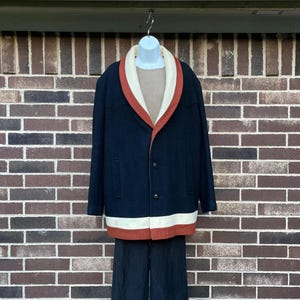 May include: A navy blue coat featuring a wide cream-colored shawl collar with a rust-orange trim. The coat has two black buttons, long sleeves, and a horizontal cream and rust-orange stripe along the bottom hem. A tan ribbed shirt is worn underneath.
