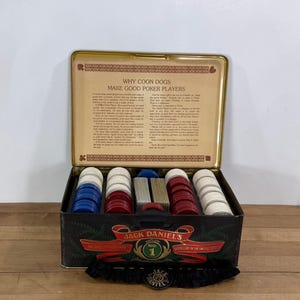 May include: A Jack Daniel's branded metal poker set case with a hinged lid featuring the text Why Coon Dogs Make Good Poker Players. The interior contains stacks of white, red, and blue poker chips and two decks of playing cards.