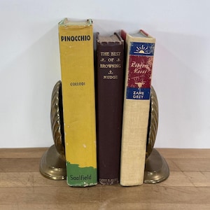 May include: A brass bookend holds three vintage books: "Pinocchio" by Collodi, "The Best of Browning" by Mudge, and "Robbers' Roost" by Zane Grey. The bookend has a seashell design.