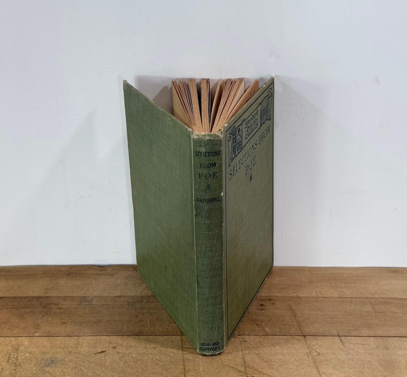 K&ouml;nnte beinhalten: Ein gebundenes Buch mit dem Titel Selections from Poe von Gambrill, herausgegeben von Ginn and Company. Das Buch hat einen verblassten olivgr&uuml;nen Stoffeinband mit dunklem Text auf dem Buchr&uuml;cken und dem Vorderdeckel. Die Seiten sind oben sichtbar und zeigen gealtertes Papier.