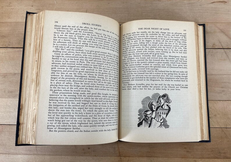 May include: An open book with aged, yellowed pages. The text is in English, with the title "Droll Stories" visible. A black and white illustration of three figures is on the right page.