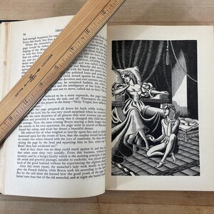 May include: An open book with text and a black and white illustration of two figures in a room. A wooden ruler rests across the pages. The book has a dark blue cover.