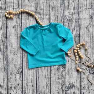 Könnte beinhalten: Ein türkisblaues Langarmshirt mit Rundhalsausschnitt. Das Shirt ist aus einem weichen Material gefertigt und wird auf einer Holzoberfläche präsentiert. Dekorative Holzperlen und Knöpfe sind um das Shirt angeordnet.