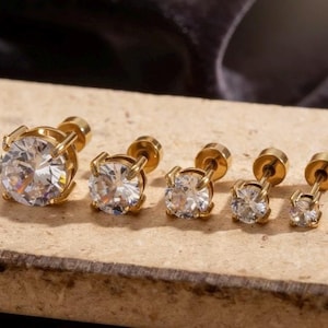 Peut inclure: Ensemble de cinq boucles d'oreilles à clous dorées avec des pierres claires, rondes et facettées. Les boucles d'oreilles sont disposées par taille, de la plus grande à la plus petite. Les pierres sont maintenues par quatre griffes.