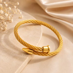 May include: A gold-toned cable wire bracelet featuring a twisted metal texture. The bracelet has an open design with two distinct ends: one end is a smooth, polished conical point, and the other is a flat, cylindrical cap with a small protruding stud.