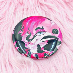 Pode incluir: Um botão redondo com um personagem de desenho animado rosa e preto estilizado com olhos azuis e cabelo rosa. O botão tem um acabamento brilhante e está sobre um fundo rosa e fofo.