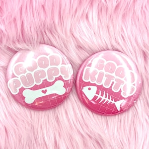 Puede incluir: Dos botones redondos rosas con texto y gráficos blancos. Uno dice "Good Puppy" con un hueso y un corazón. El otro dice "Good Kitty" con un diseño de espina de pescado. Los botones están sobre una superficie rosa y esponjosa.