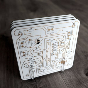 May include: A white circuit board with gold-coloured traces and components. The board is mounted on a clear acrylic stand. The board has labels for various components, including resistors, capacitors, and integrated circuits.