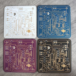 Könnte beinhalten: Vier quadratische Untersetzer mit einem Schaltkreisdesign. Die Untersetzer sind weiß, blau, lila und schwarz mit goldfarbenen Schaltkreisen.