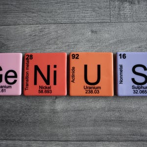 May include: Four colorful square tiles spelling out the word "Genius" with the chemical symbol and atomic number for each element. The tiles are pink, orange, orange, and purple.